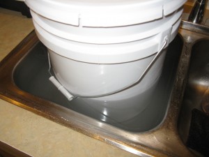 A 5 gallon bucket of crystallized/solidified honey, resting inside a sinkfull of hot water to help liquefy the honey.